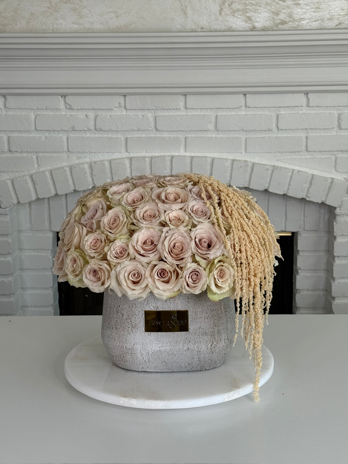 A floral arrangement of blush pink roses in a textured gray pot with dried beige grasses draping from the sides, displayed on a round white marble stand in front of a white brick fireplace.
