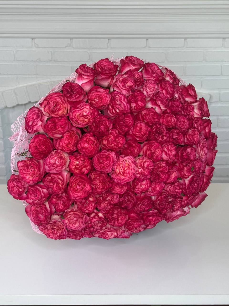 A large, dense bouquet of vibrant magenta-pink roses arranged in a heart-like shape, wrapped in light pink lace, sitting on a white surface against a white brick wall background.