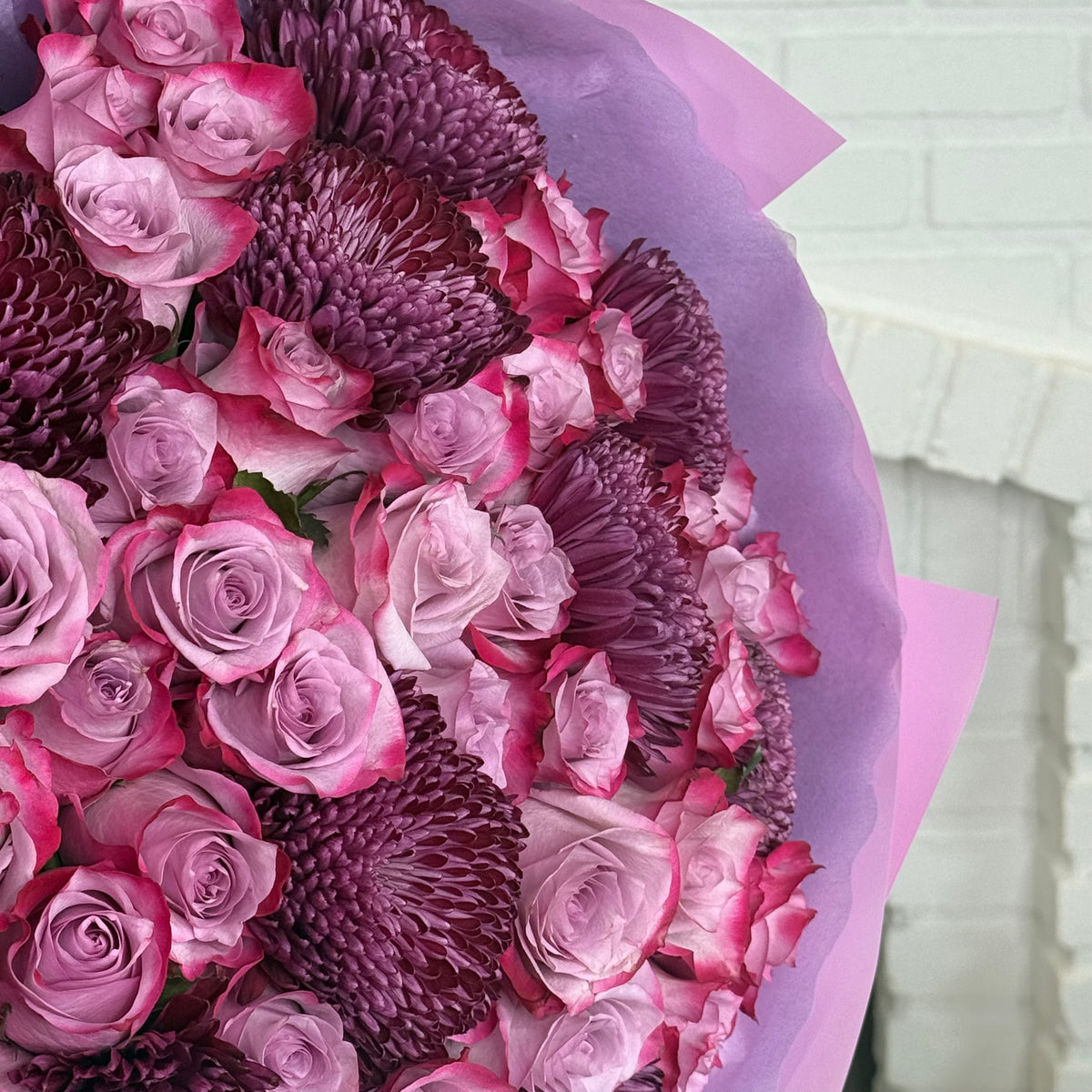 A large floral bouquet filled with pink and purple roses and protea-like flowers, wrapped in light purple tissue paper, photographed against a white brick wall background.