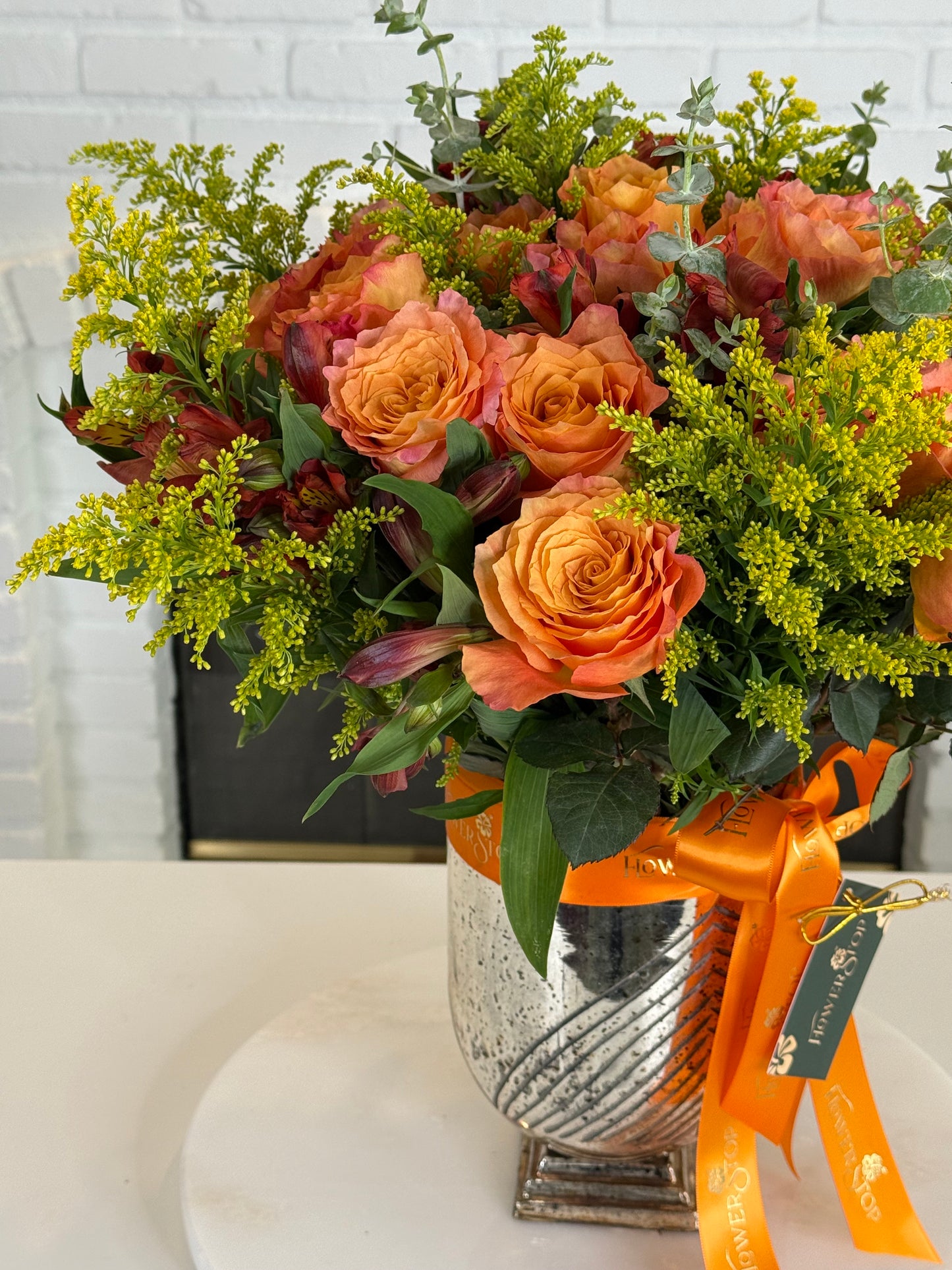 A vibrant autumn floral arrangement in a textured silver vase, featuring orange roses, yellow-green filler flowers, and green foliage, tied with an orange ribbon.