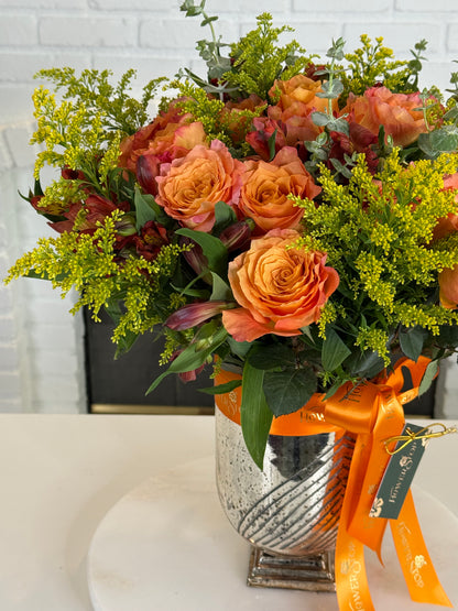 A vibrant autumn floral arrangement in a textured silver vase, featuring orange roses, yellow-green filler flowers, and green foliage, tied with an orange ribbon.