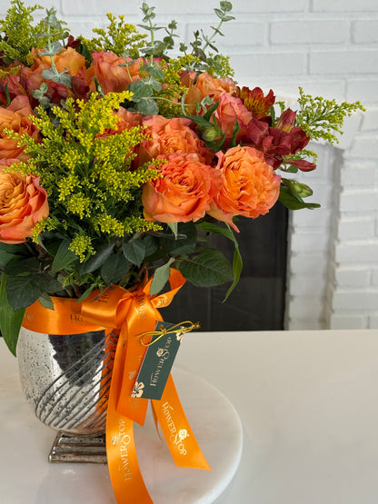 A vibrant autumn floral arrangement in a silver speckled vase, featuring orange roses, yellow filler flowers, and green foliage, tied with an orange ribbon and a green 'Flowers Direct' tag, set on a white surface against a white brick wall background.