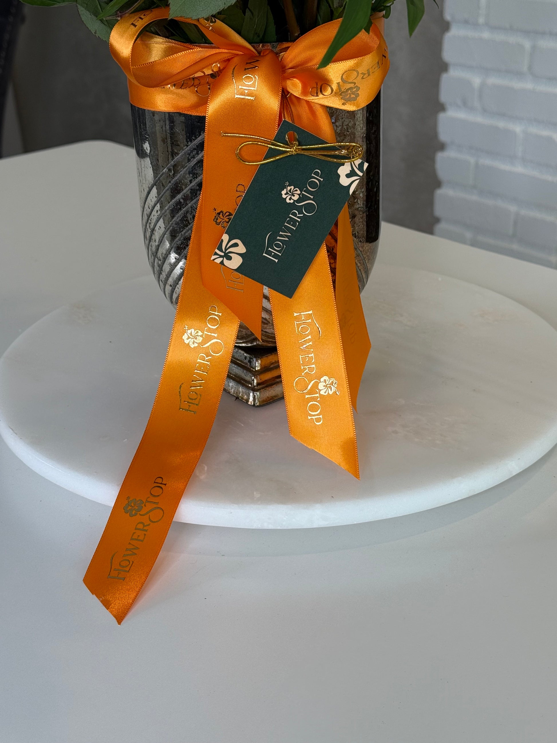 A close-up of a silver metallic vase wrapped with a bright orange ribbon and a dark green hanging tag printed with the Flower Shop name, displayed on a white round pedestal.