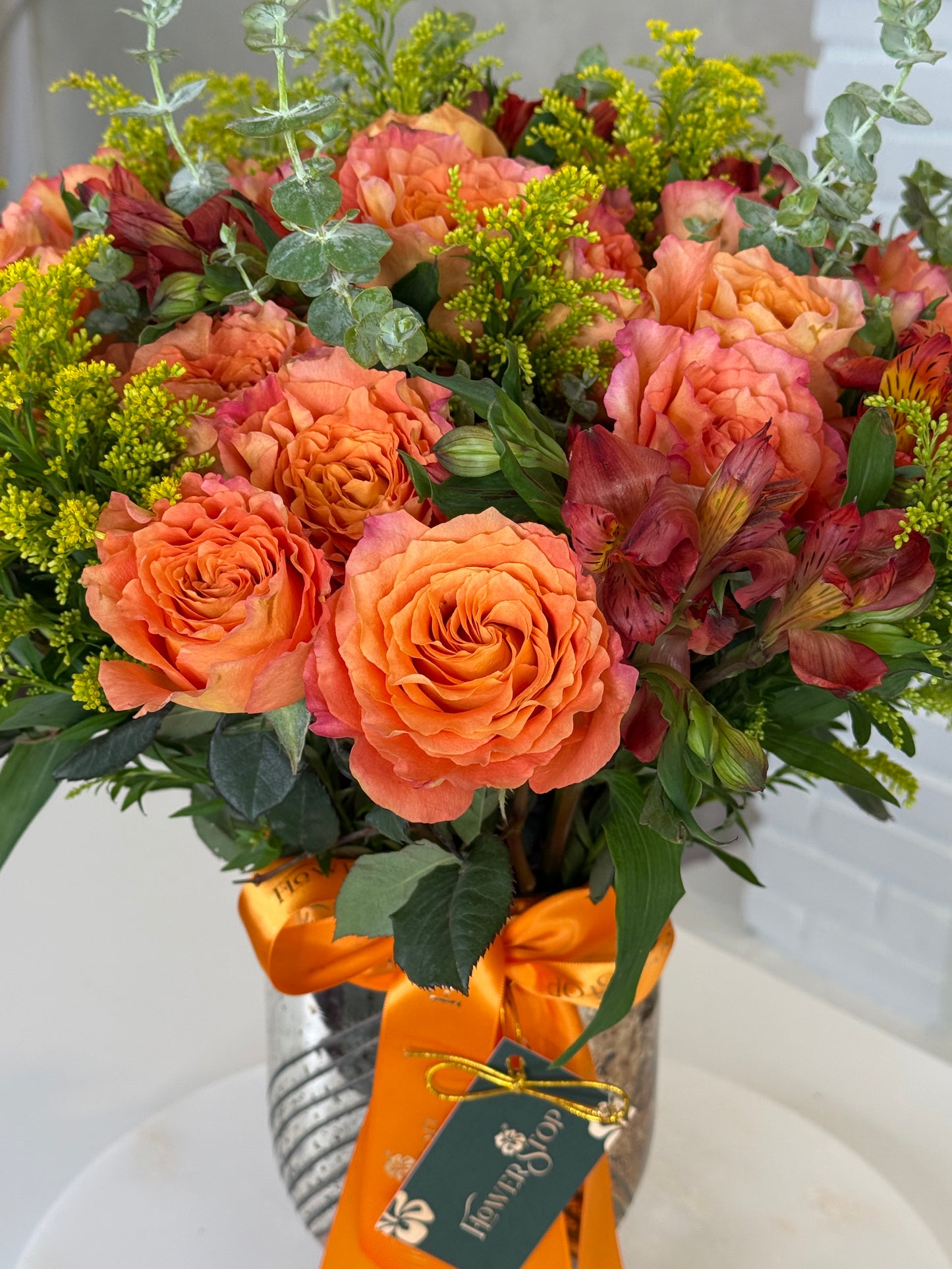 A vibrant floral arrangement featuring orange roses, peach alstroemeria, and green foliage in a silver pot with an orange ribbon and a 'Flower Box' gift tag.