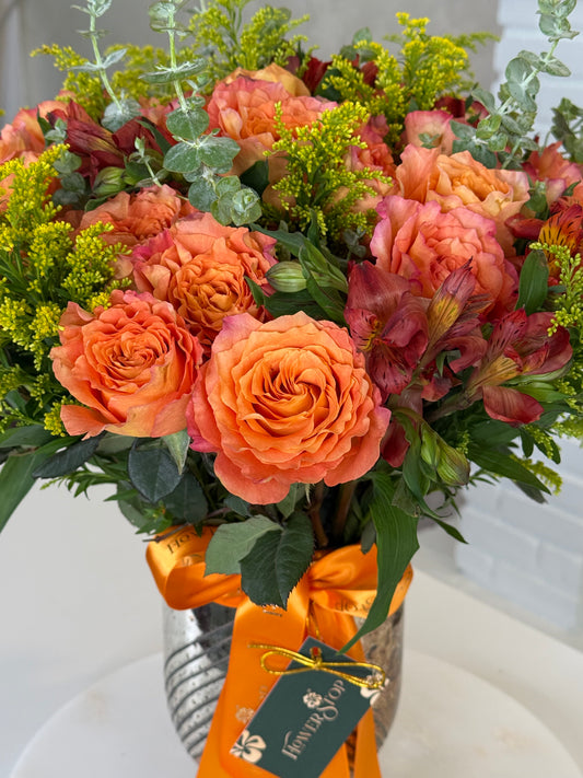 A vibrant floral arrangement featuring orange roses, peach alstroemeria, and green foliage in a silver pot with an orange ribbon and a 'Flower Box' gift tag.