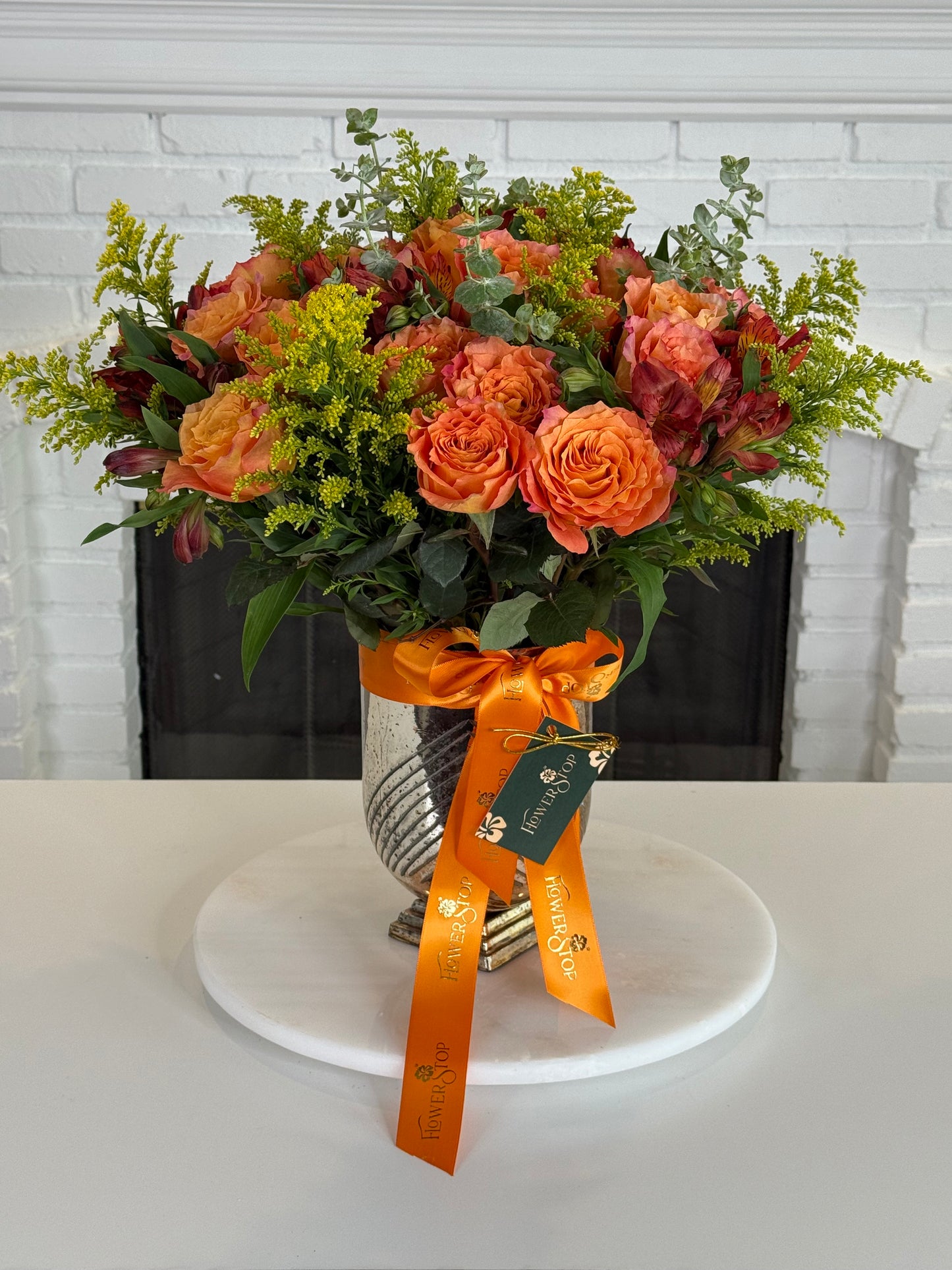 An autumn floral arrangement featuring orange roses, burgundy dahlias, and green foliage in a textured silver vase, tied with an orange ribbon and displayed on a white marble platter in front of a white brick fireplace.