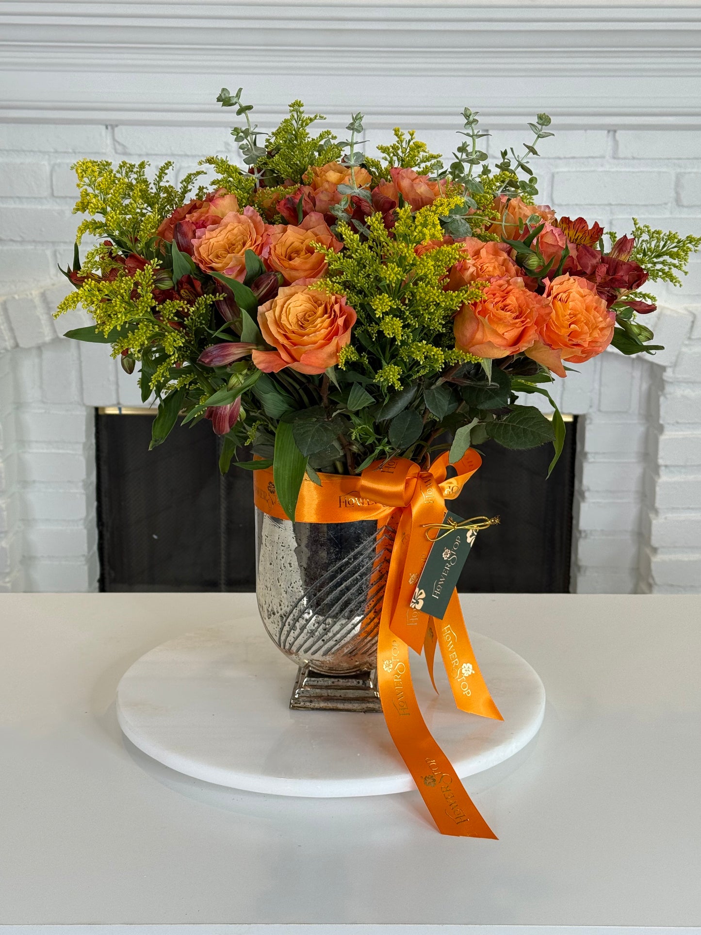 An autumn floral arrangement in a textured silver vase, featuring orange roses, yellow filler flowers, and green foliage, tied with an orange ribbon on a white round platter in front of a white brick fireplace.