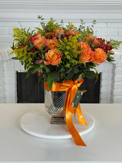 An autumn floral arrangement in a textured silver vase, featuring orange roses, yellow filler flowers, and green foliage, tied with an orange ribbon on a white round platter in front of a white brick fireplace.