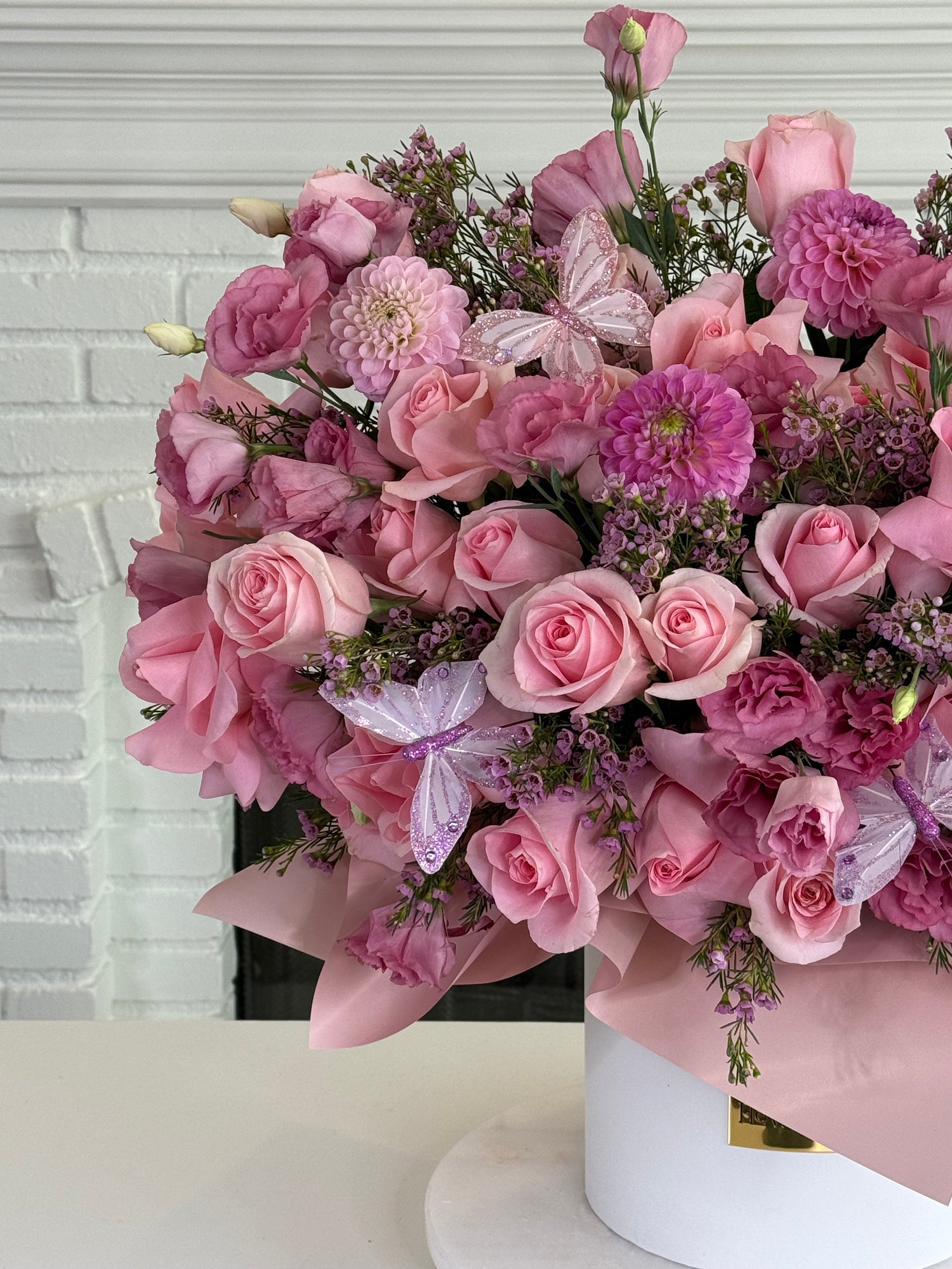 A large, vibrant pink floral arrangement featuring roses, chrysanthemums, and butterfly flowers, wrapped in light pink paper and presented in a white cylindrical pot in front of a white brick fireplace.