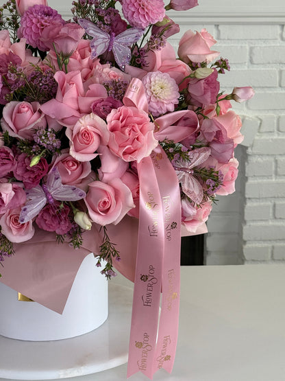A large, lush pink floral arrangement in a white round box, featuring pink roses, carnations, hydrangeas, and decorative purple butterflies, tied with a pink branded ribbon.