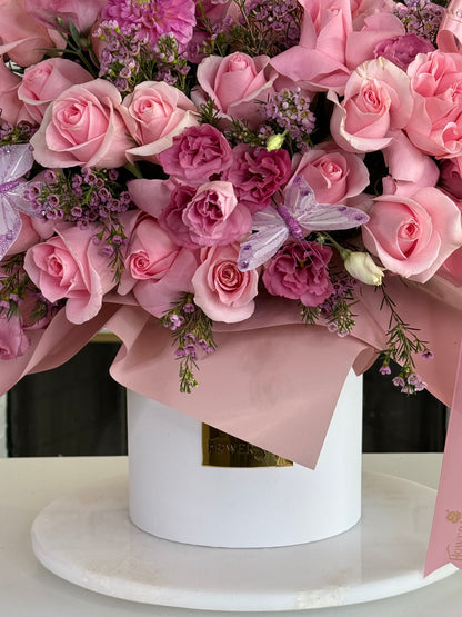 A pink floral arrangement featuring pink and magenta roses, purple filler flowers, and a purple dragonfly ornament, presented in a white cylindrical box wrapped with blush pink paper on a white marble surface.