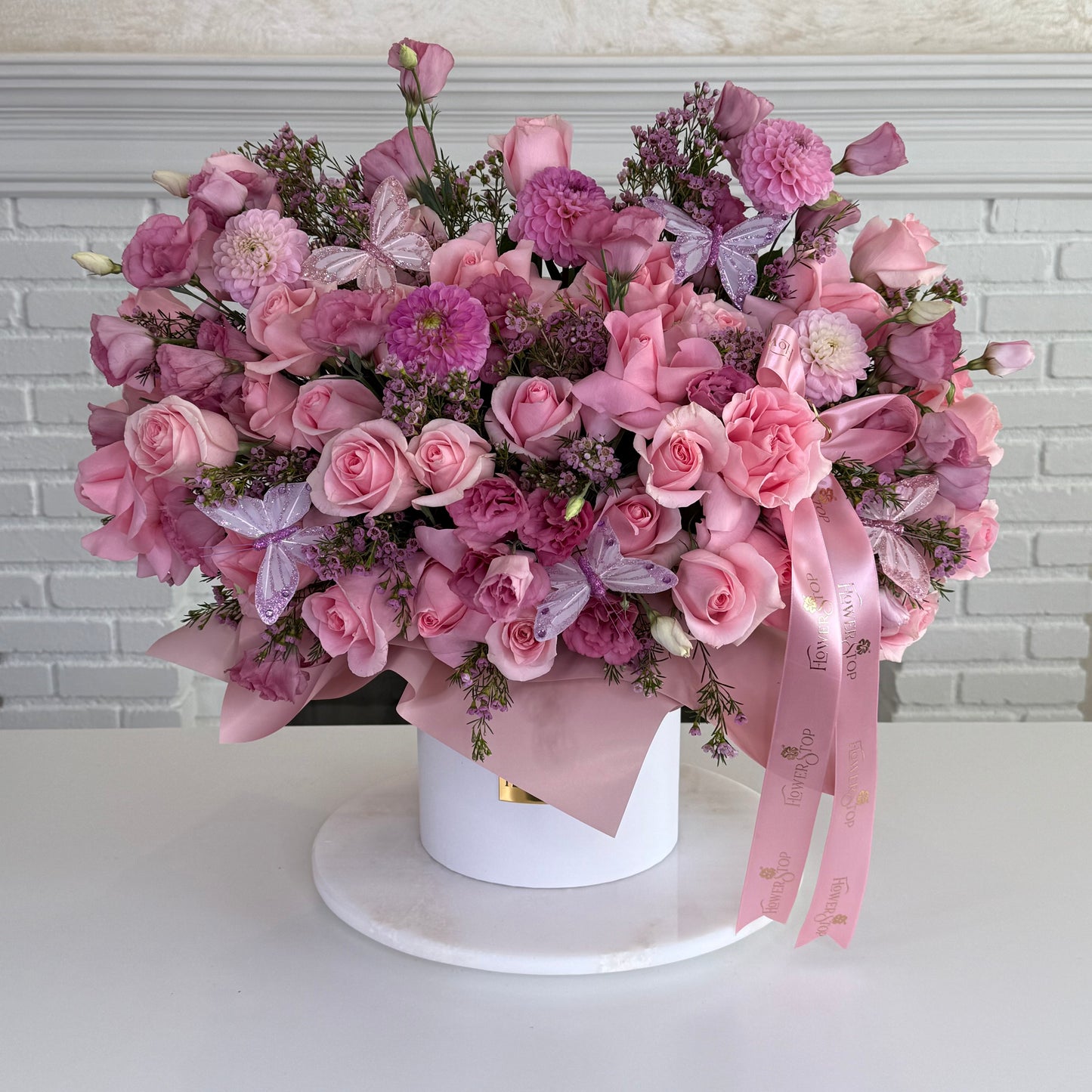 A large, vibrant pink floral arrangement featuring roses, chrysanthemums, and butterfly flowers, styled in a white round box with a pink ribbon, set on a white marble coaster against a white brick wall background.