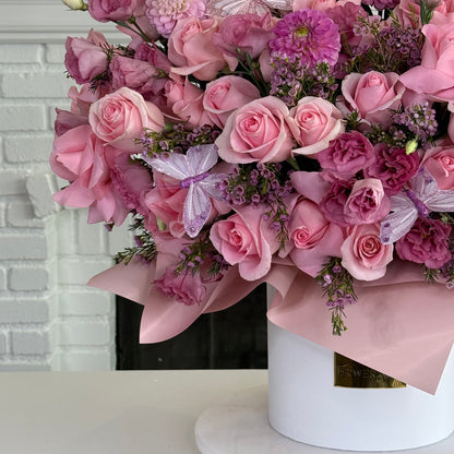 A large floral arrangement of pink roses, pink carnations, and purple filler flowers with butterfly embellishments, wrapped in light pink paper and presented in a white cylindrical box on a white surface.