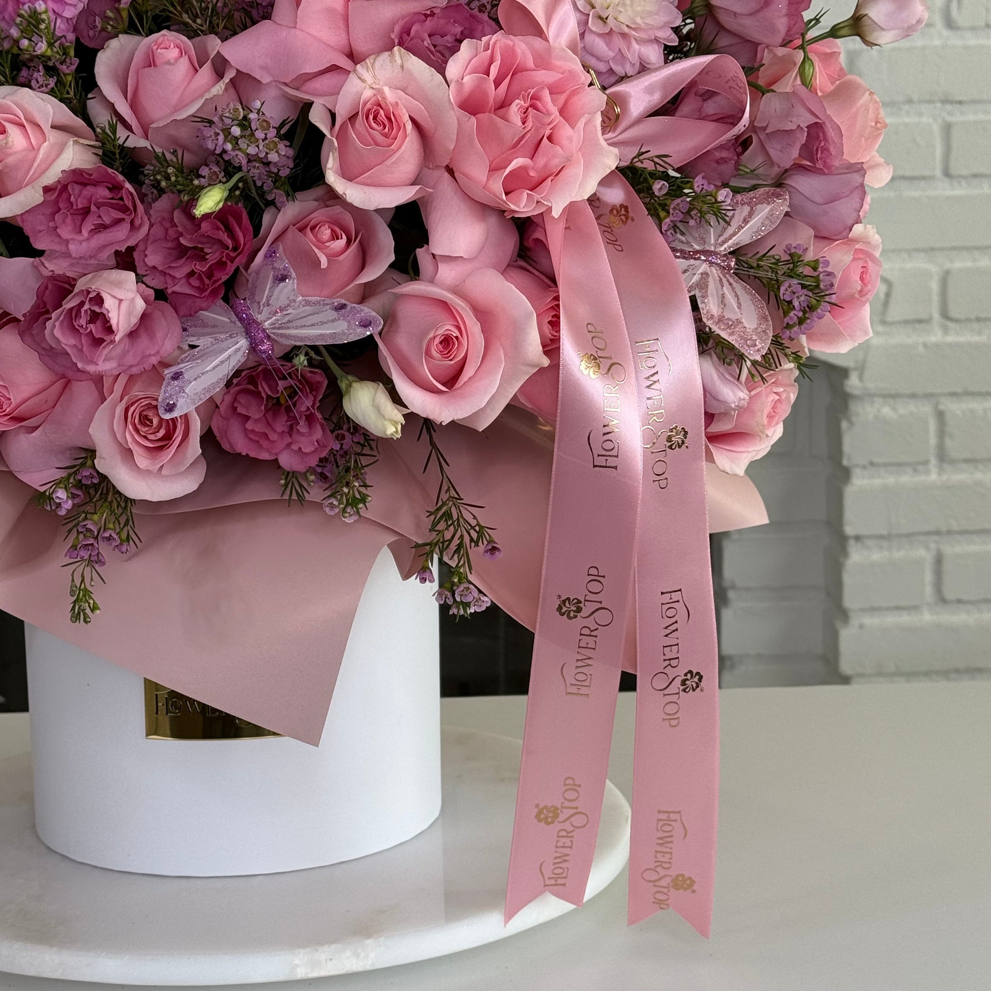 A large pink floral arrangement in a white cylindrical box, featuring pink roses, purple carnations, and a pink ribbon printed with the Flower Shop logo, set on a white surface against a light gray brick wall.