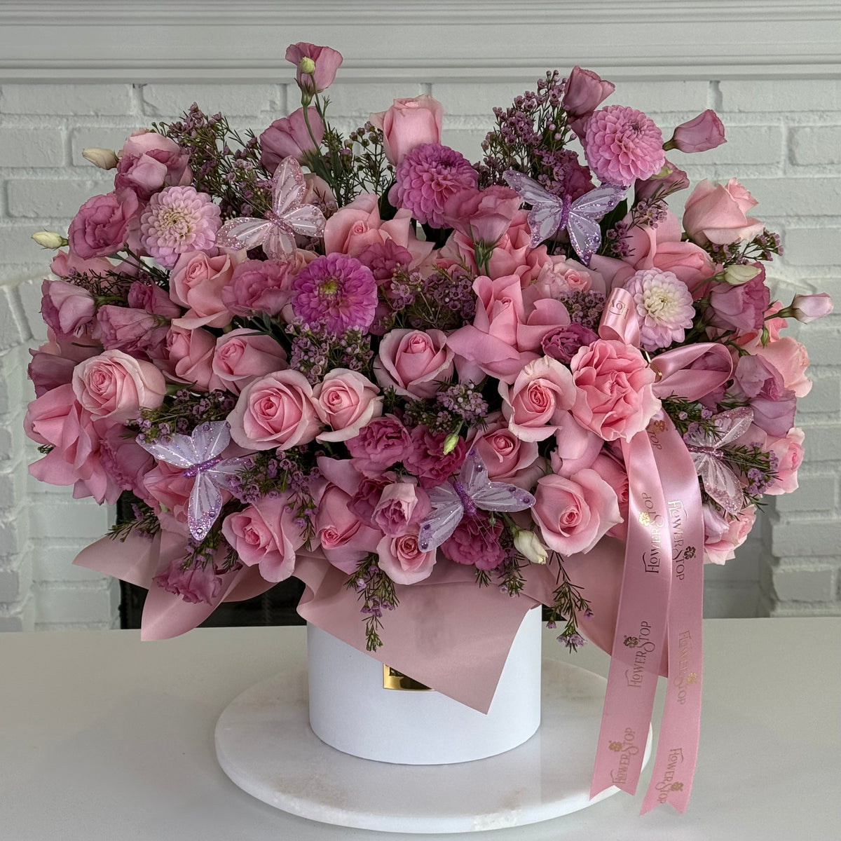 A large, dense floral arrangement in a white cylindrical box, featuring pink and purple roses, chrysanthemums, and butterfly flowers, wrapped in pink paper with a pink ribbon, set on a white round pedestal against a white brick wall background.