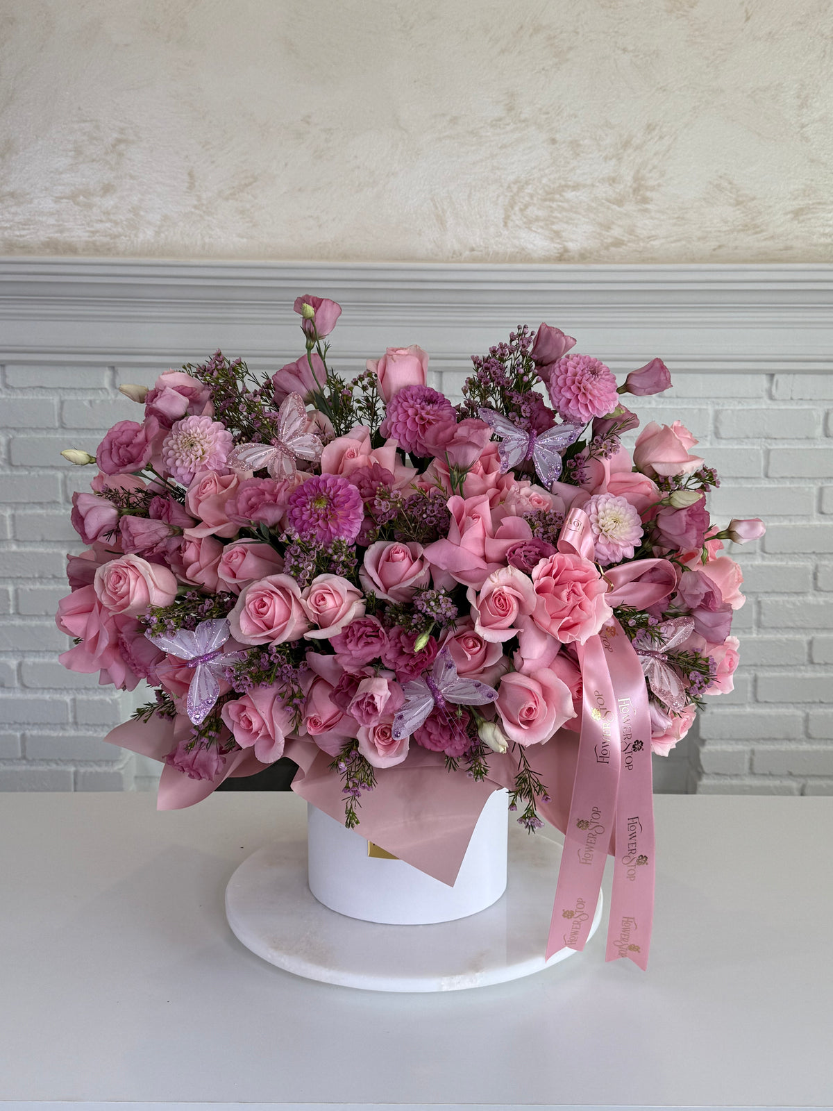 A large heart-shaped floral arrangement in shades of pink and purple, presented in a white cylindrical box with a pink ribbon, set on a white table against a white brick wall background.
