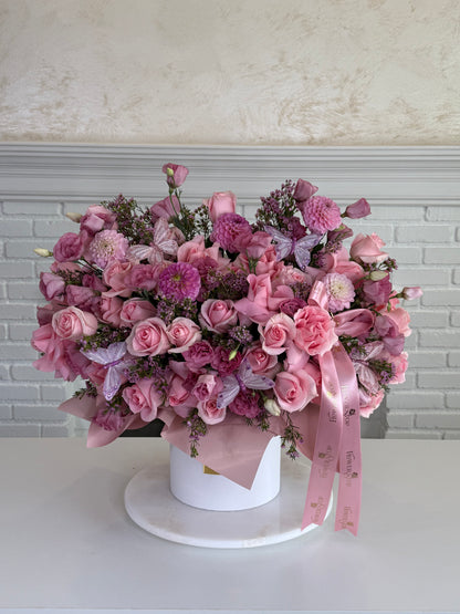 A large heart-shaped floral arrangement in shades of pink and purple, presented in a white cylindrical box with a pink ribbon, set on a white table against a white brick wall background.