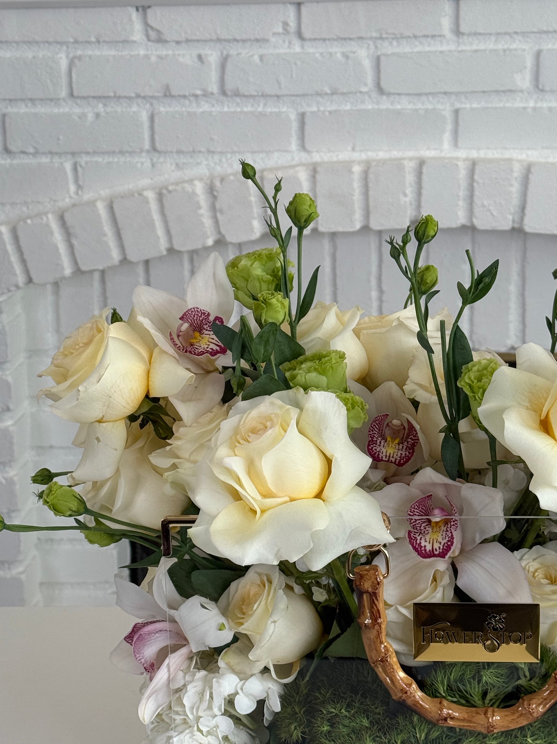 A floral arrangement featuring cream and white roses, white orchids with pink centers, and green foliage in a woven basket, set against a white brick wall background.