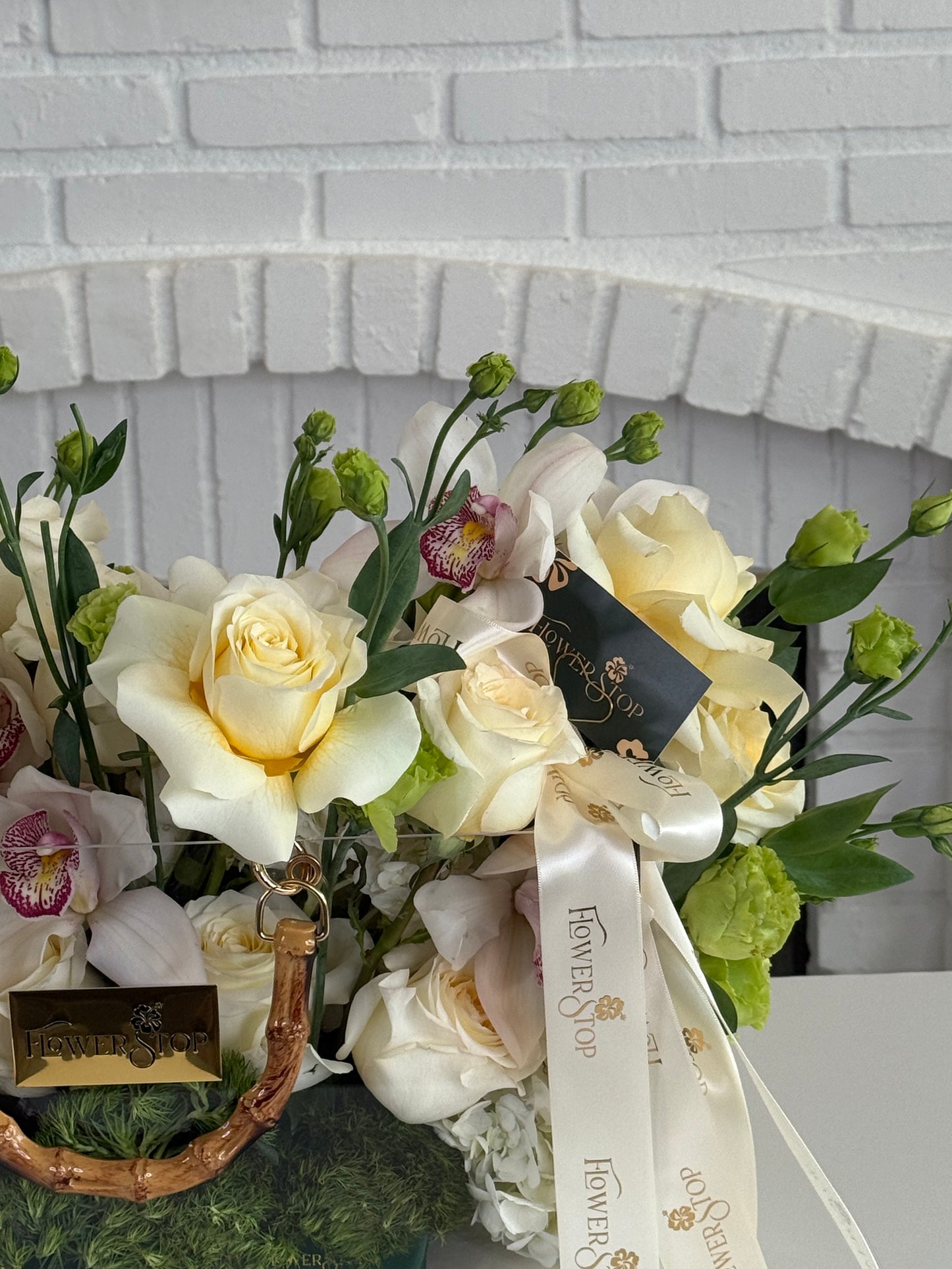 A floral arrangement featuring cream and pale yellow roses, green buds, and orchids in a woven basket, tied with a white ribbon printed with the brand name 'Flower of the Year'.