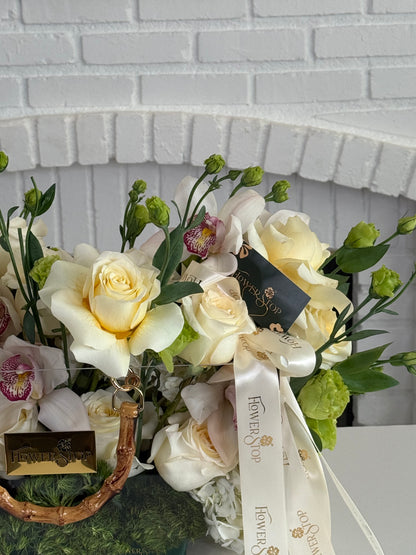 A floral arrangement featuring cream and pale yellow roses, green buds, and orchids in a woven basket, tied with a white ribbon printed with the brand name 'Flower of the Year'.