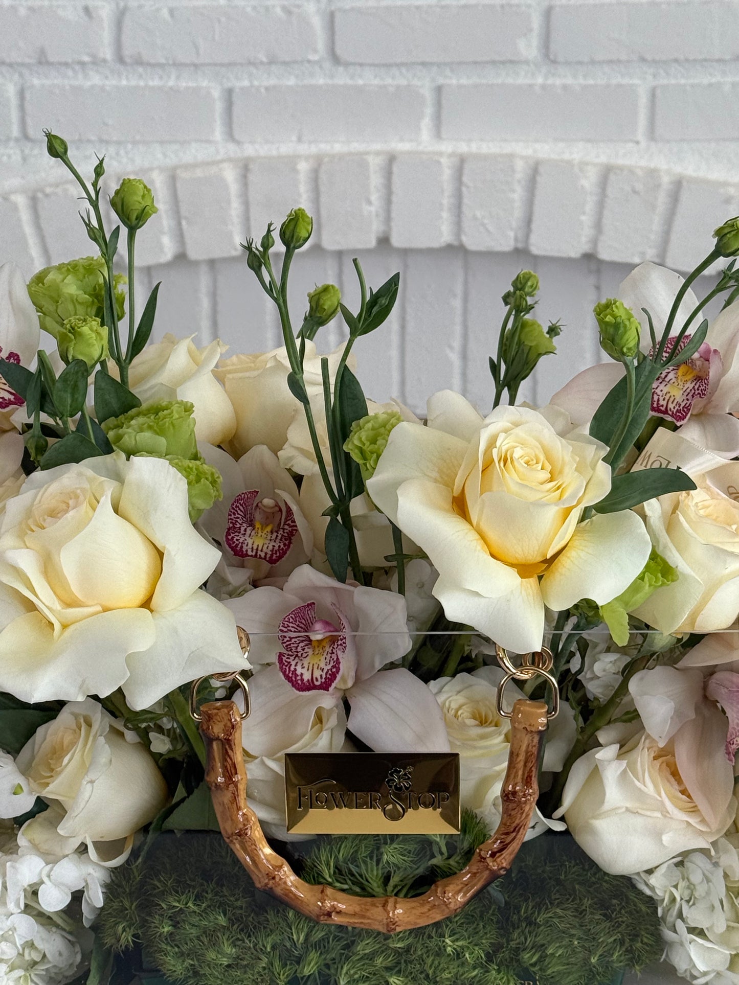 A floral arrangement featuring cream and white roses, pale orchids with pink centers, green buds, and green foliage in a basket with a bamboo handle and a gold-tone brand plaque reading 'HENDRETS'.