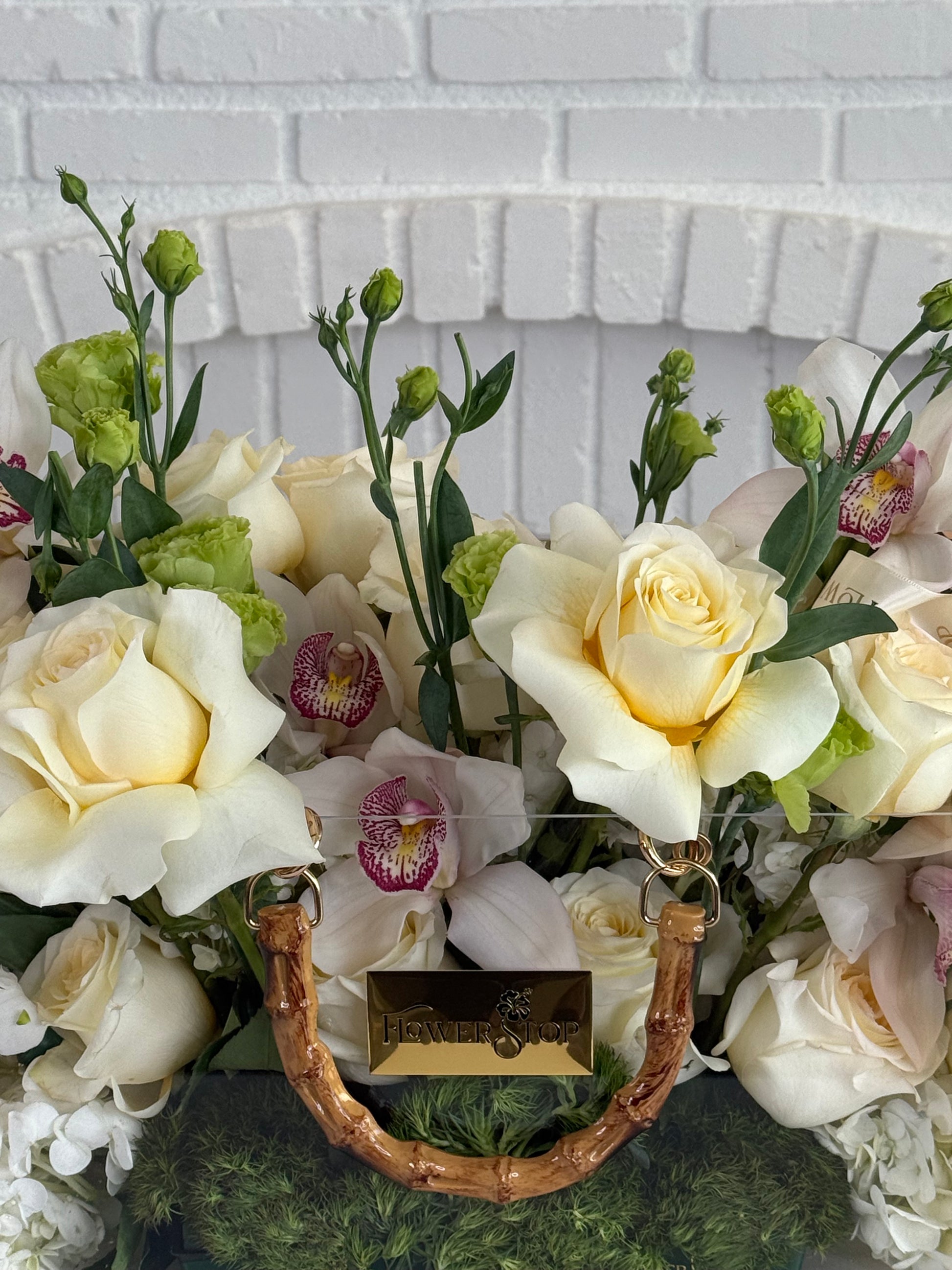 A floral arrangement featuring cream and white roses, pale orchids with pink centers, green buds, and green foliage in a basket with a bamboo handle and a gold-tone brand plaque reading 'HENDRETS'.