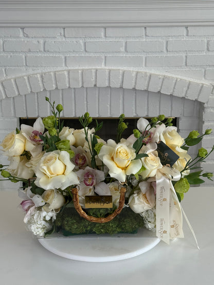 A floral arrangement in a clear rectangular vase with a gold handle, featuring cream roses, white orchids, green foliage, and moss, set against a white brick fireplace. A cream ribbon with gold lettering is tied to the side.