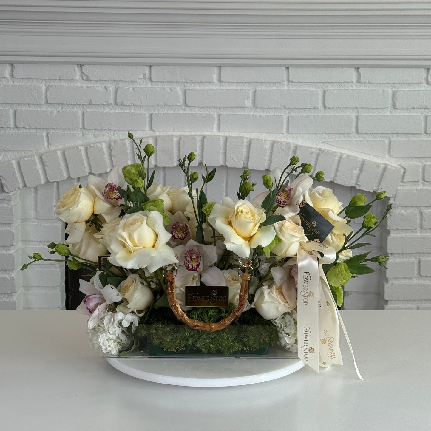 A floral arrangement of cream and white roses, green foliage, and small pink blossoms in a woven basket with a gold handle, displayed on a white round stand in front of a white brick fireplace.