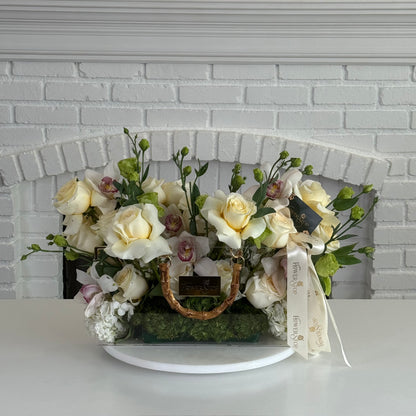 A floral arrangement of cream and white roses, green foliage, and small pink blossoms in a woven basket with a gold handle, displayed on a white round stand in front of a white brick fireplace.