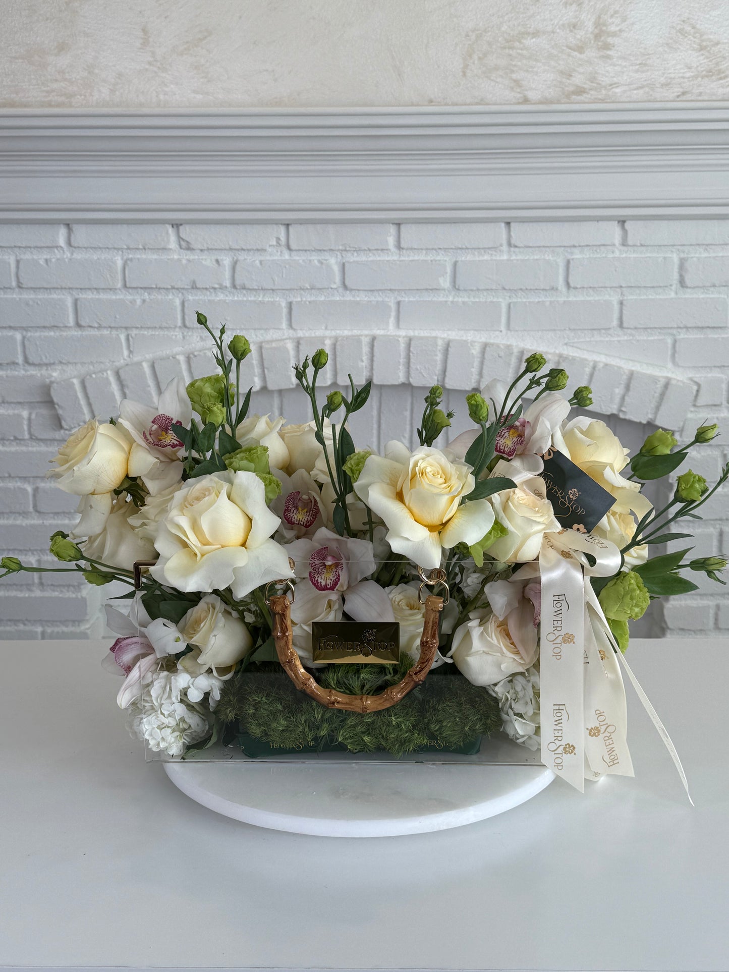 A floral arrangement in a clear rectangular planter with a wicker-style handle, featuring cream and pale yellow roses, white orchids, green foliage, moss, and a white ribbon, displayed on a white pedestal against a white brick wall.