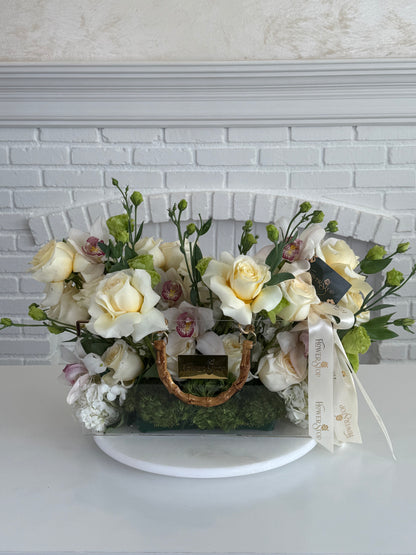 A floral arrangement in a clear rectangular planter with a wicker-style handle, featuring cream and pale yellow roses, white orchids, green foliage, moss, and a white ribbon, displayed on a white pedestal against a white brick wall.