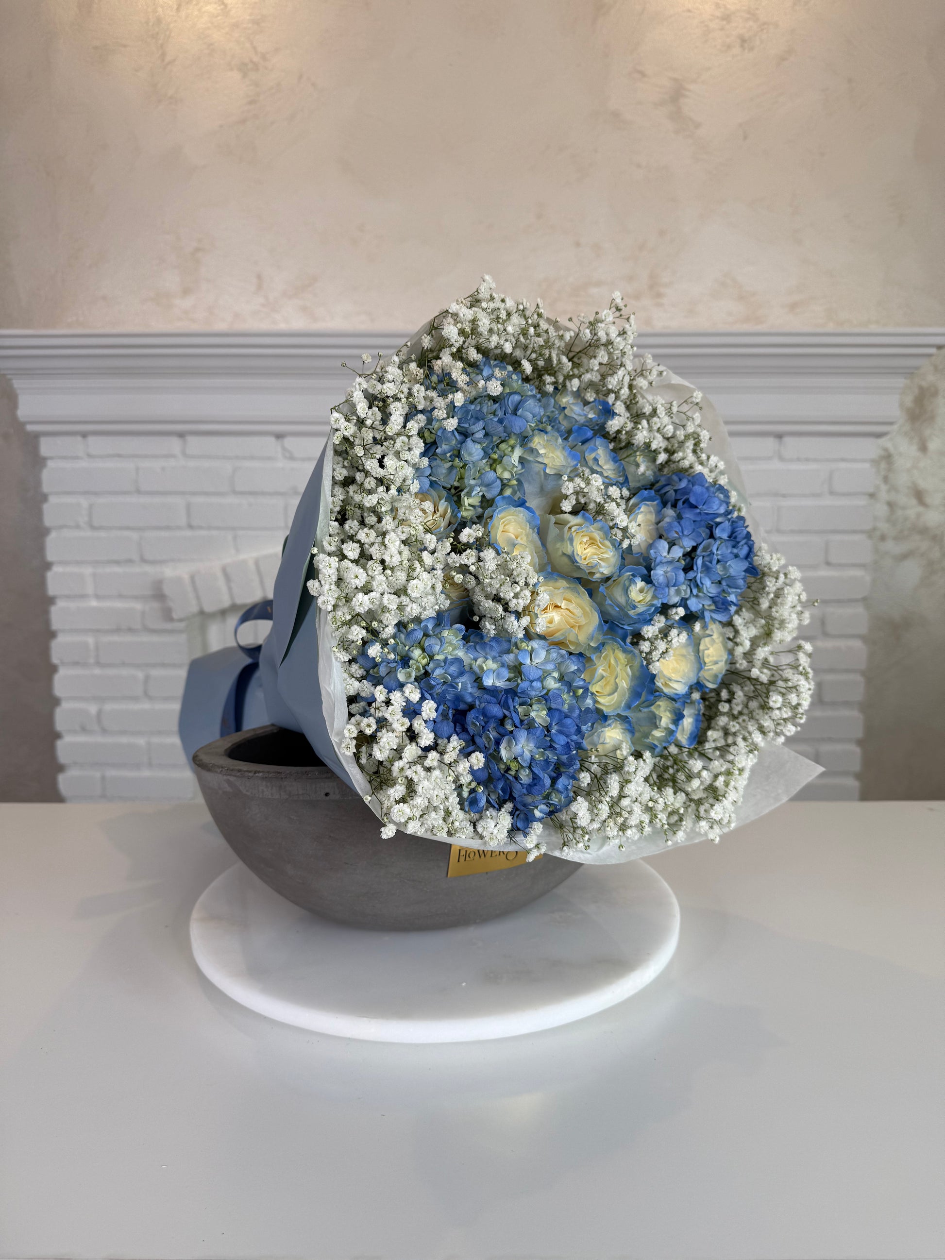 A dried flower bouquet featuring blue hydrangeas, pale yellow roses, and white baby's breath, wrapped in a blue-gray paper sleeve and displayed on a white circular stand.