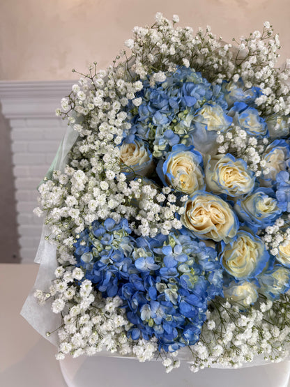 A bouquet of blue and cream roses mixed with blue hydrangeas and abundant white baby's breath, wrapped in white tissue paper.