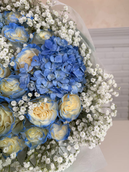 A bouquet of blue and cream roses mixed with blue hydrangeas and white baby's breath, wrapped in light gray paper.
