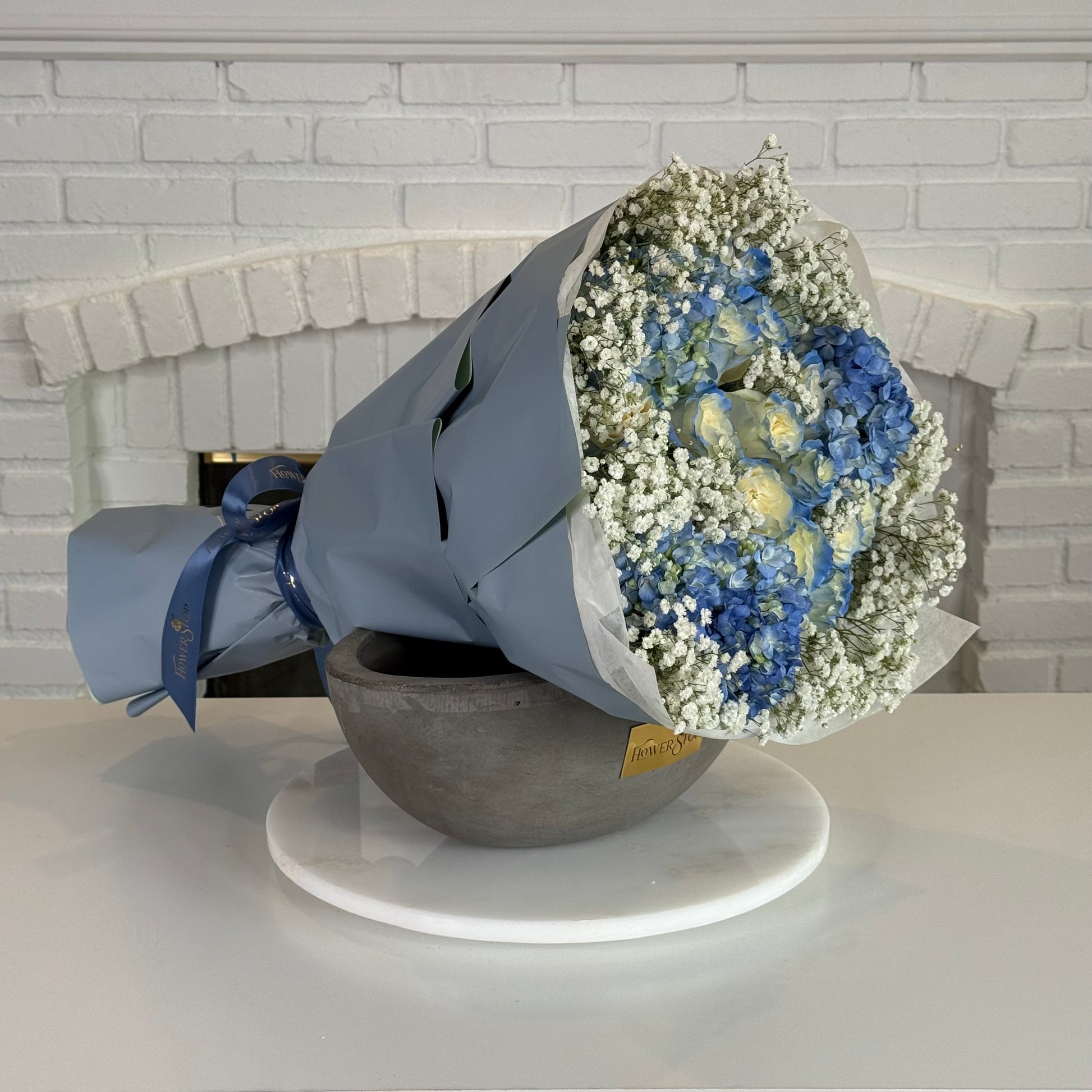 A wrapped flower bouquet with blue hydrangeas, white baby's breath, and round yellow flowers, displayed in a gray bowl on a white round tray against a white brick fireplace background.