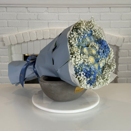 A wrapped flower bouquet with blue hydrangeas, white baby's breath, and round yellow flowers, displayed in a gray bowl on a white round tray against a white brick fireplace background.