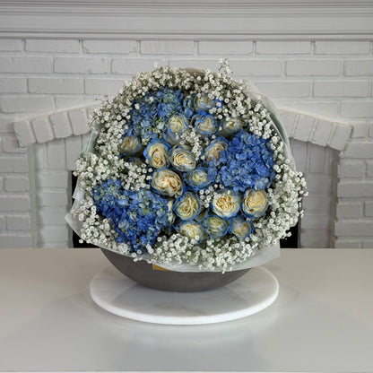 A round floral arrangement of blue hydrangeas and pale yellow roses with white baby's breath, displayed in a gray bowl on a white round stand against a white brick wall background.