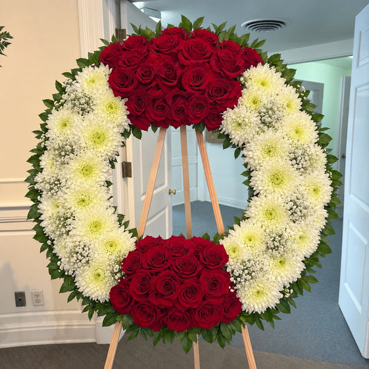 A large circular floral wreath displayed on a wooden easel stand in an indoor hallway. The wreath features red roses at the top and bottom center, surrounded by white chrysanthemums and small white baby's breath with green foliage.