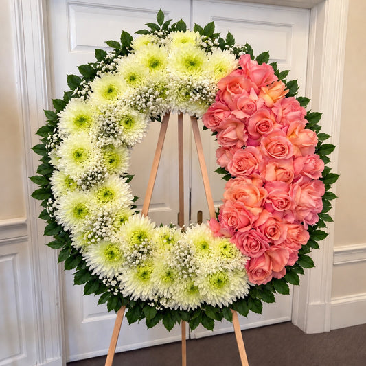A large circular floral wreath displayed on a wooden stand in front of white paneled doors. The wreath is divided into two halves: one side features white chrysanthemums with green centers and small white baby's breath, while the other side features pink and peach roses with green leaves.