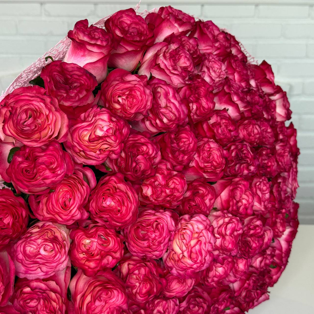 A large round bouquet of vibrant magenta and pink roses, tightly packed and wrapped in sheer pink paper, set against a white brick wall background.