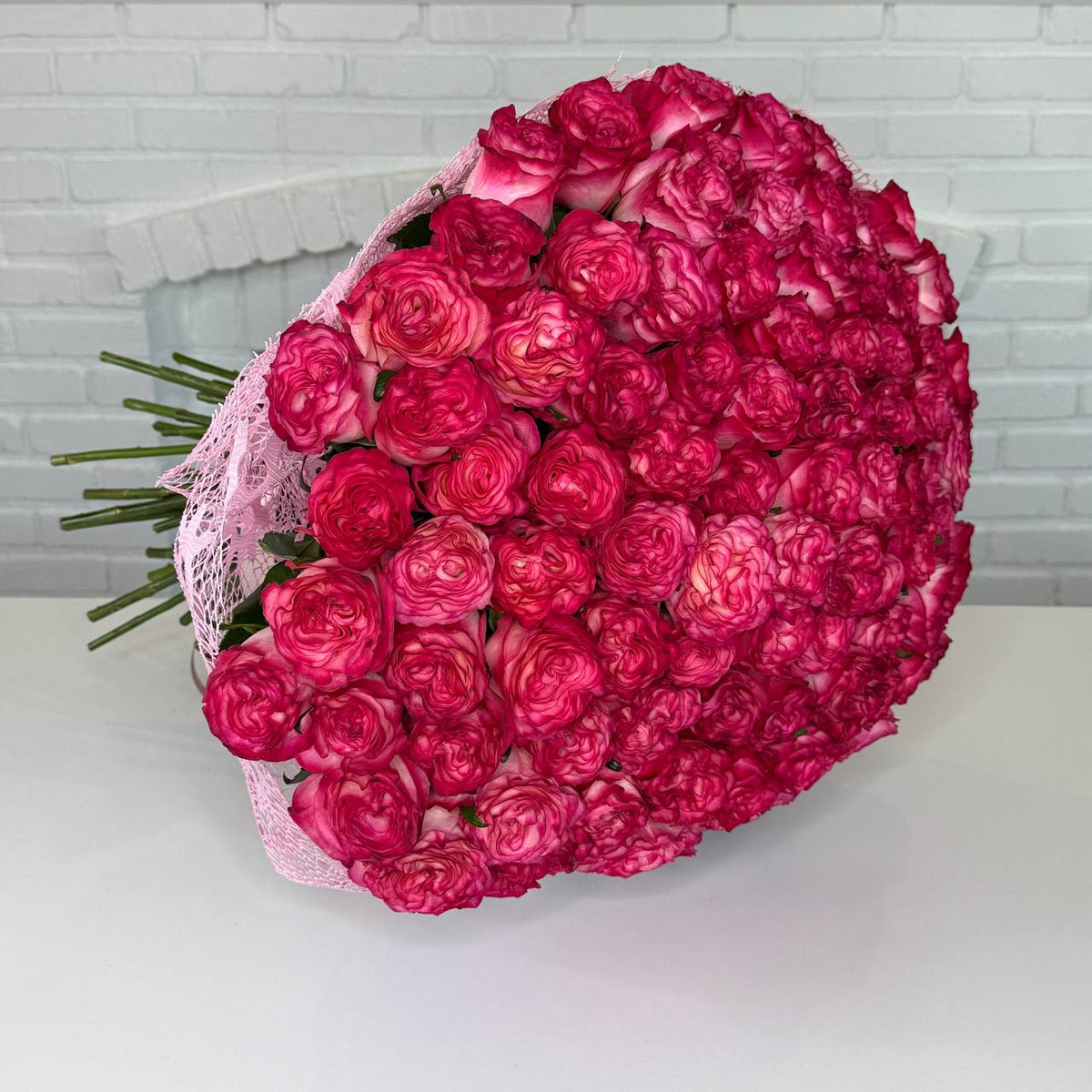 A large bouquet of vibrant pink roses wrapped in light pink lace, displayed on a white table against a white brick wall background.