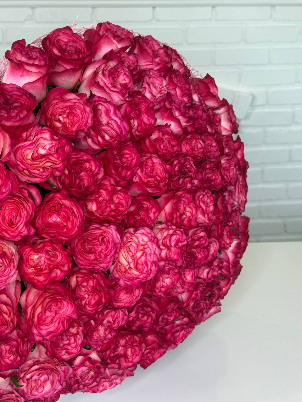 A large, heart-shaped bouquet of vibrant magenta-pink roses arranged on a white surface against a white brick wall background.