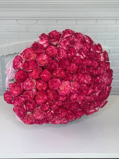 A large, dense bouquet of vibrant magenta-pink roses arranged in a heart-like shape, wrapped in light pink lace, sitting on a white surface against a white brick wall background.