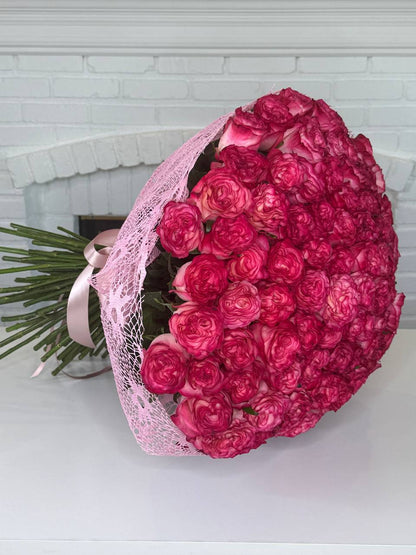 A large bouquet of deep pink roses wrapped in light pink lace and tied with a matching pink ribbon, placed on a white surface in front of a white brick fireplace.