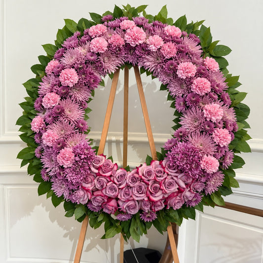 A circular floral wreath displayed on a wooden stand, featuring pink and purple flowers including carnations, chrysanthemums, and roses, with green leaves throughout, set against a white paneled wall.