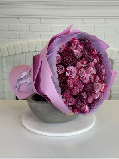 A bouquet of pink and purple flowers wrapped in lavender paper, displayed in a gray bowl on a white round stand against a white brick wall background.