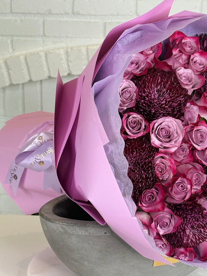 A bouquet of pink and purple flowers wrapped in lavender paper and tied with a matching ribbon, displayed in a gray bowl against a white brick wall background.