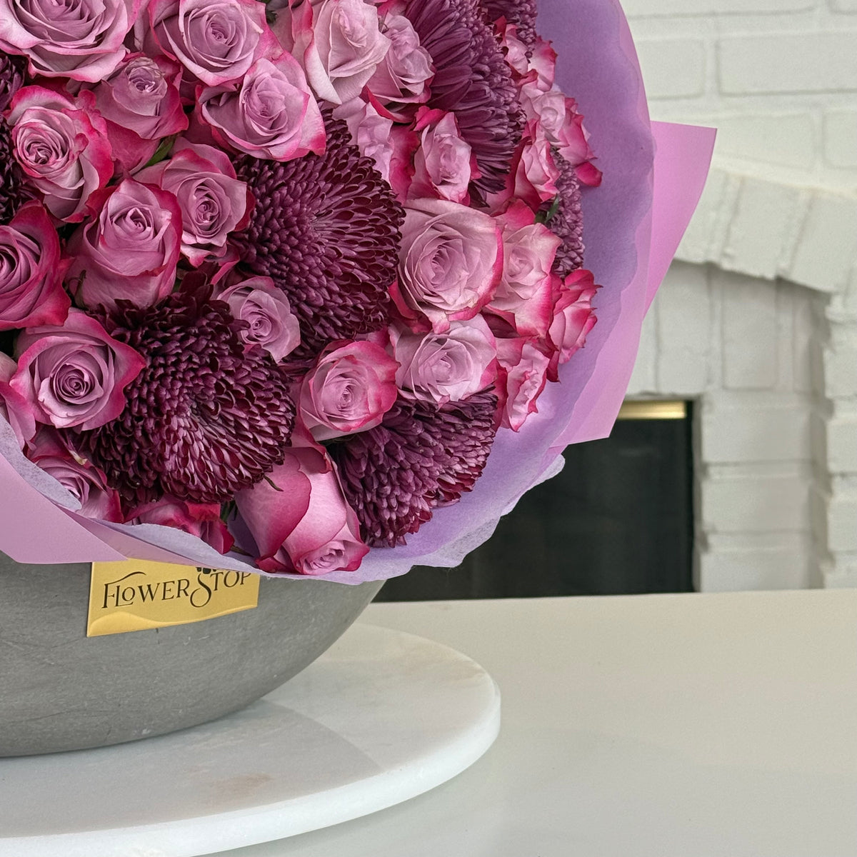A large floral bouquet in a gray bowl with a lavender wrapper and a small Flower Stop label, photographed on a white surface with a white brick fireplace in the background.