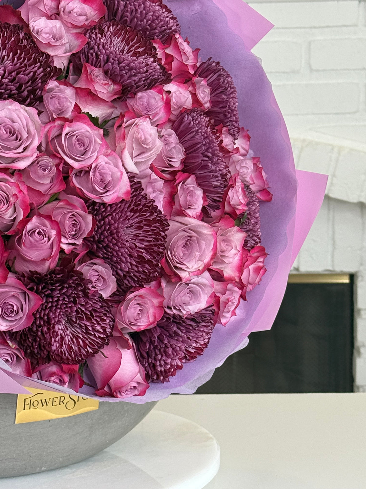 A large floral bouquet featuring pink and purple roses mixed with deep magenta chrysanthemums, wrapped in lavender paper and placed on a white surface with a white brick wall in the background.