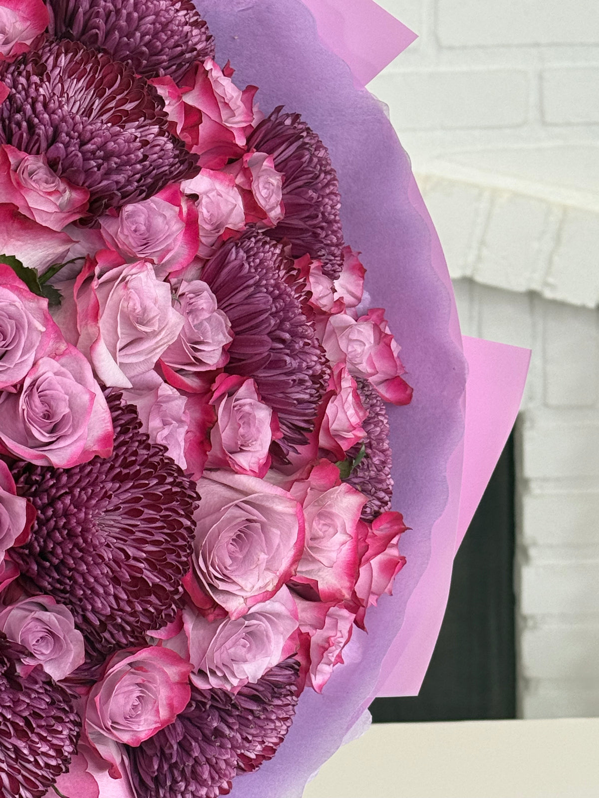 A close-up of a floral bouquet wrapped in light purple tissue paper, featuring pink and purple roses and textured chrysanthemums, set against a white brick wall background.